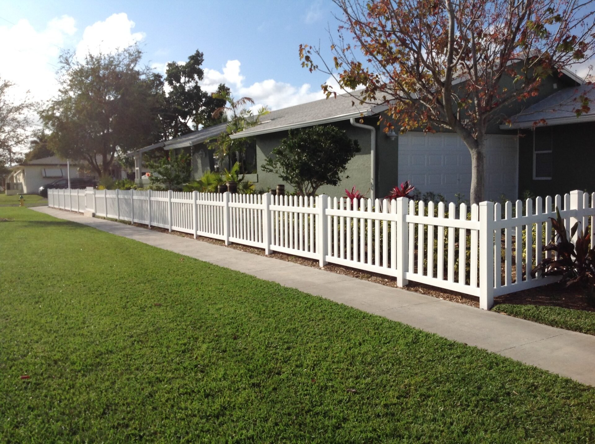 A white picket style vinyl fence encloses the yard of a ranch house with a maple tree.