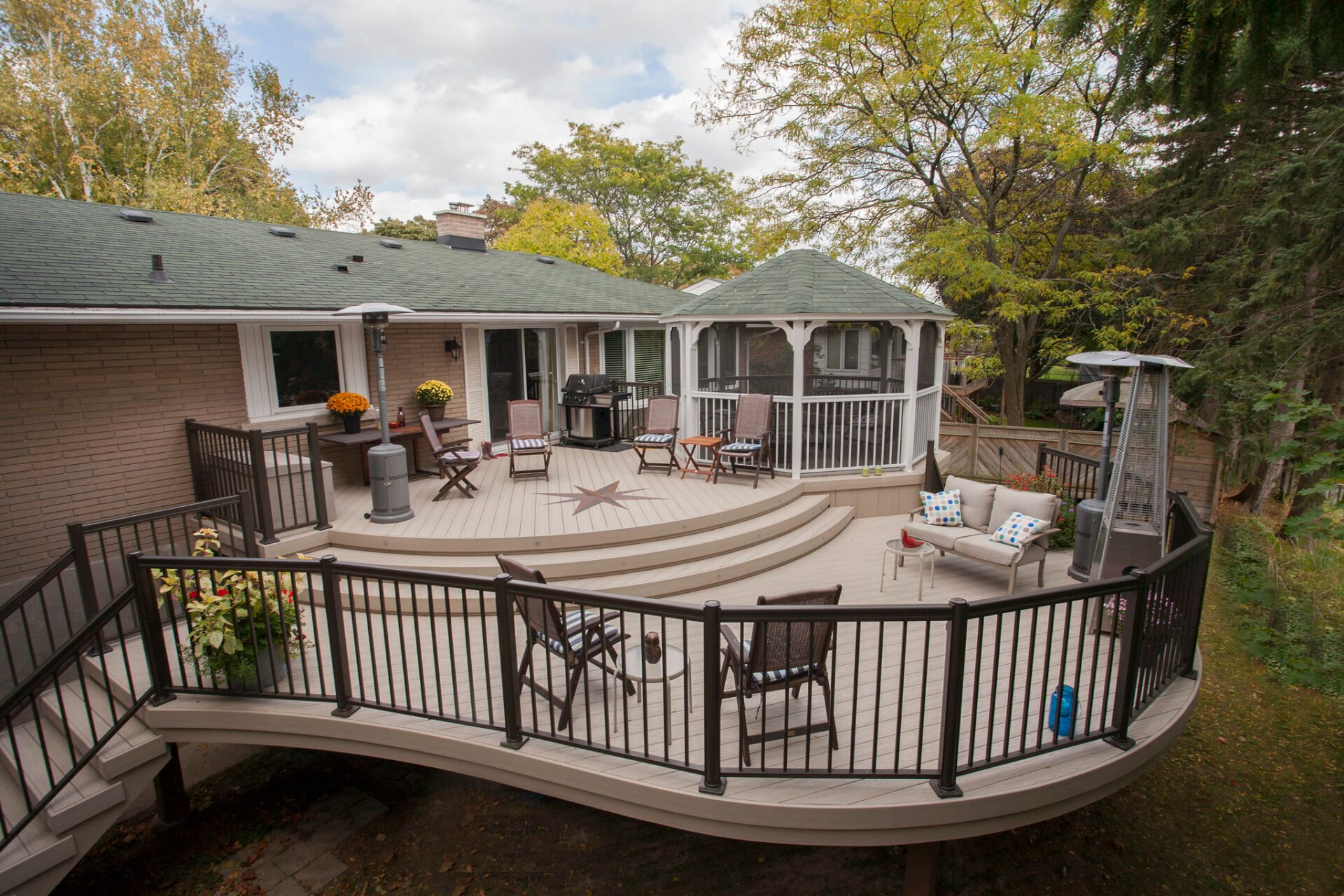 A multilevel custom deck with beige decking, black aluminum rails, and a gazebo, attached to a tan brick house.