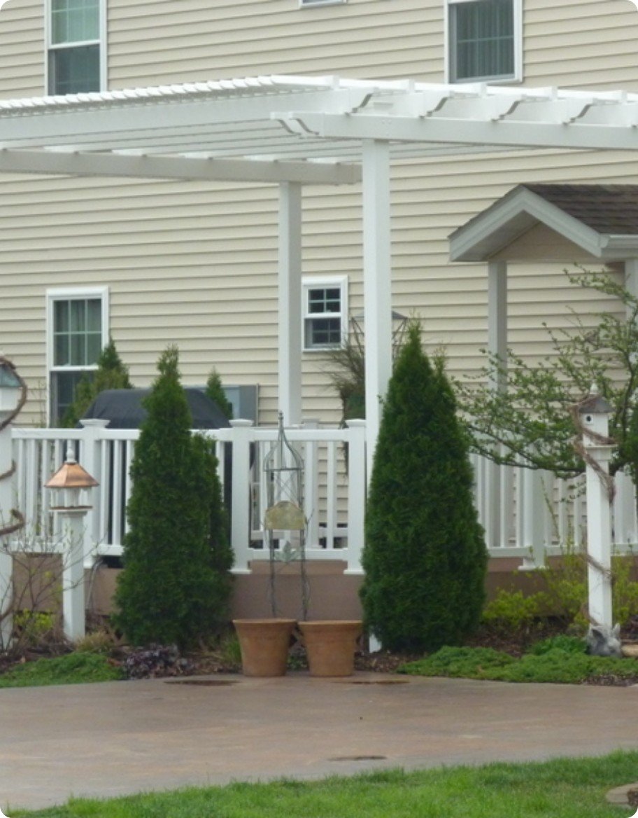 A deck with white vinyl railings and a white vinyl pergola behind a beige home.