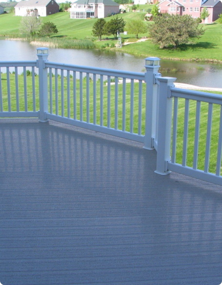 A gray deck with white vinyl railing overlooking a sunny backyard and pond.