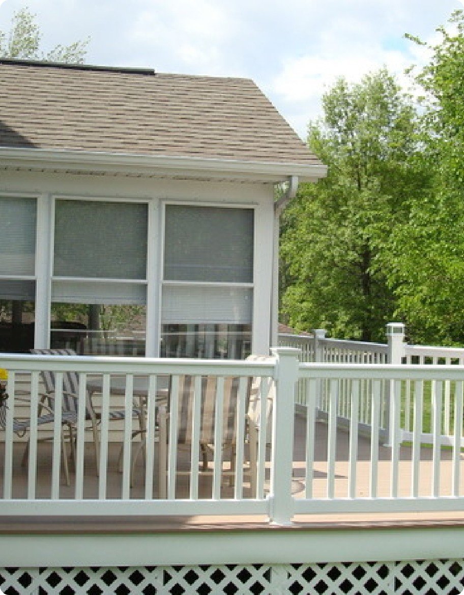 A brown deck in a wooded backyard with white vinyl railings and latticework sides.