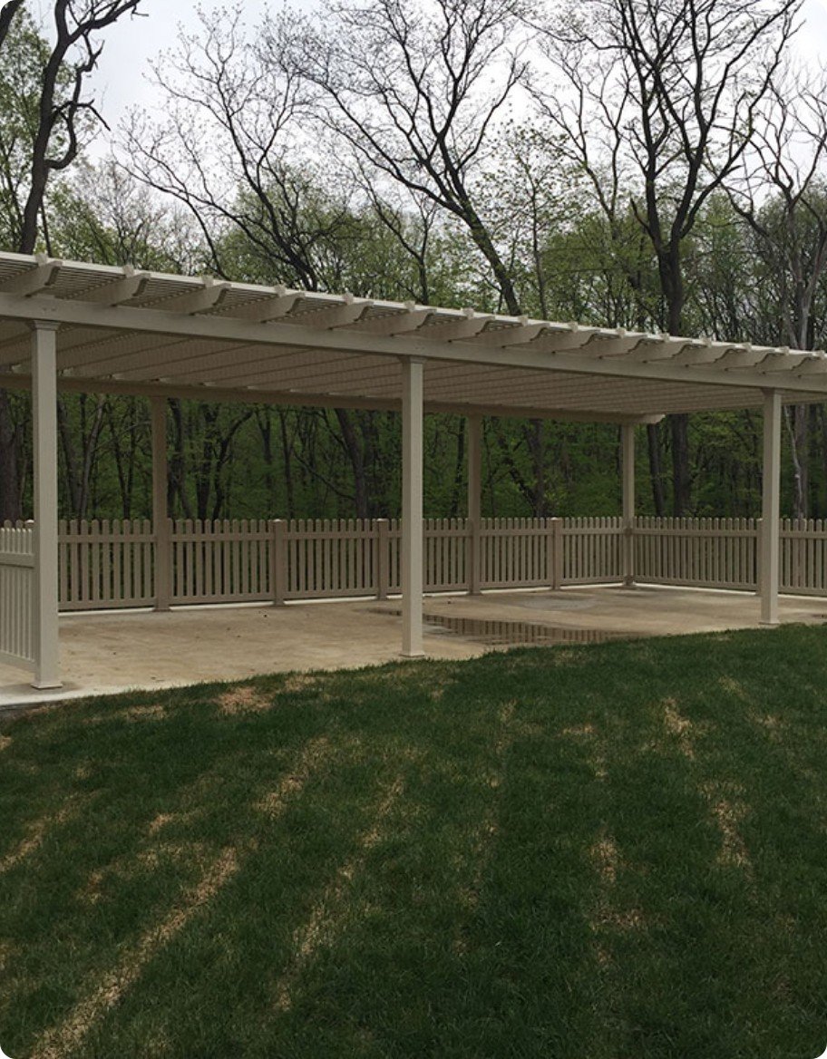 A large tan vinyl pergola over a patio surrounded by a tan picket fence.