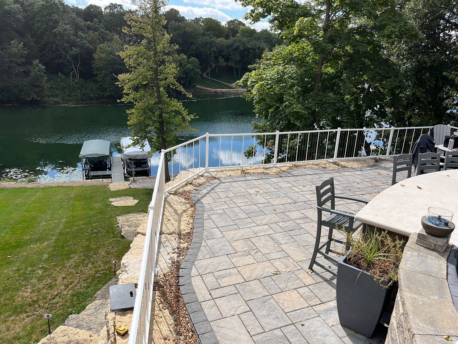 A stone-tiled deck with white cable railings overlooks a peaceful river and two docked boats.