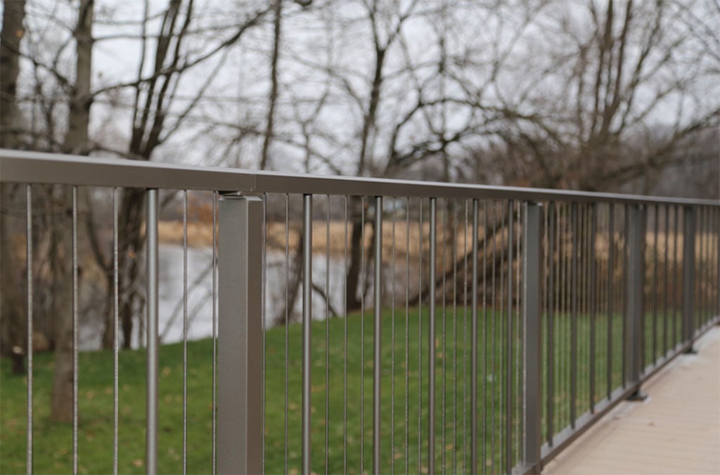 A view through a gray aluminum railing with vertical cable supports toward a pond.