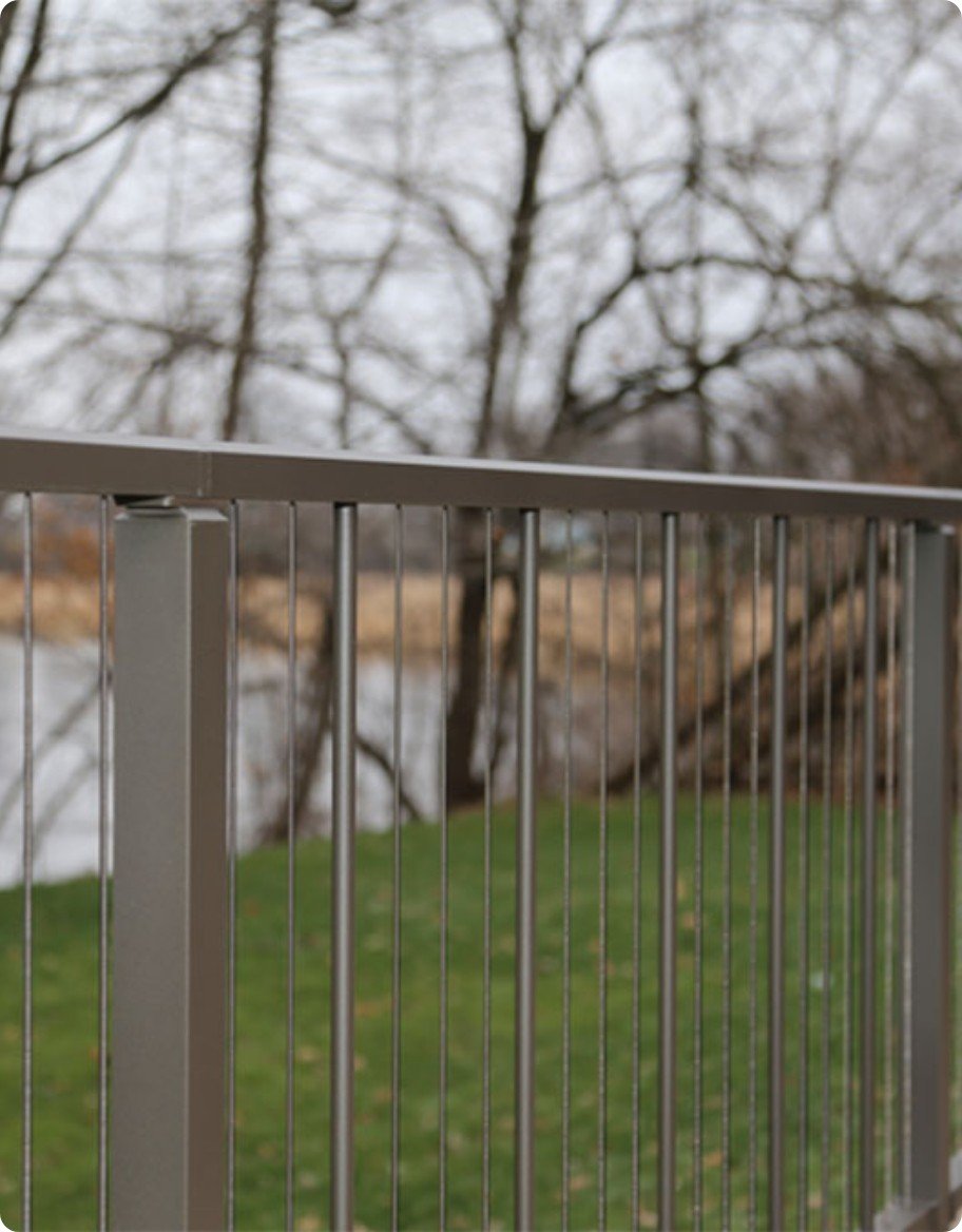 A gray aluminum deck railing with vertical cable supports, looking toward a pond.