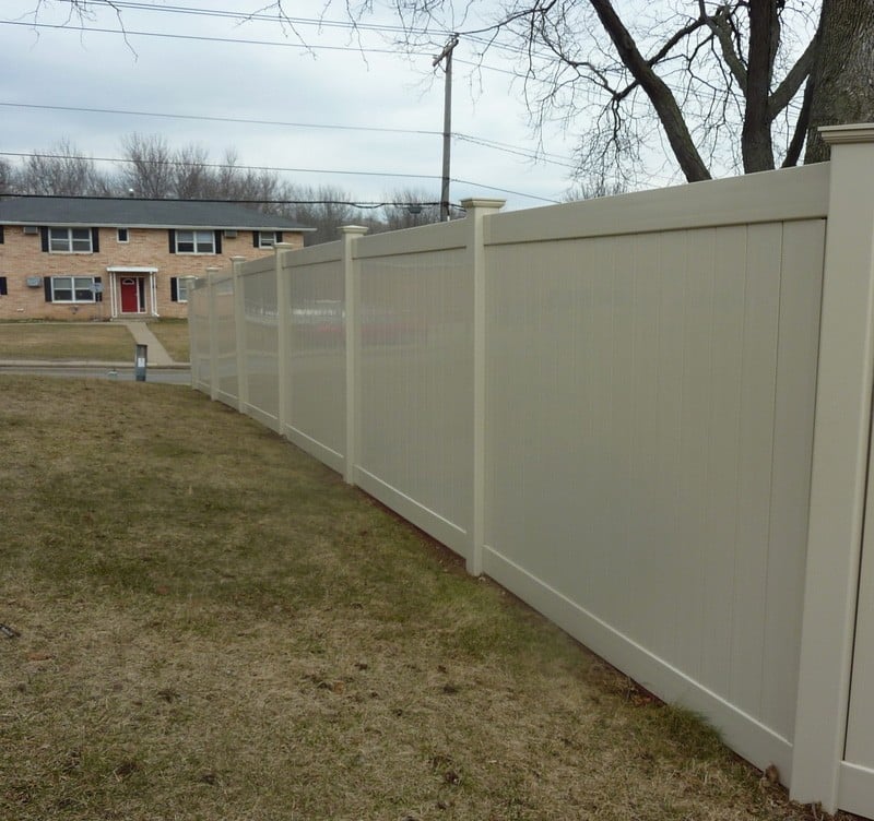 A tall vinyl privacy fence in a beige color, with a tan brick apartment house across the street.