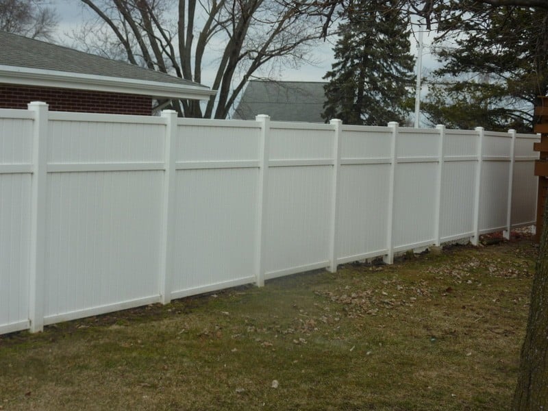 A tall white vinyl privacy fence borders a well-kept yard on an autumn day.