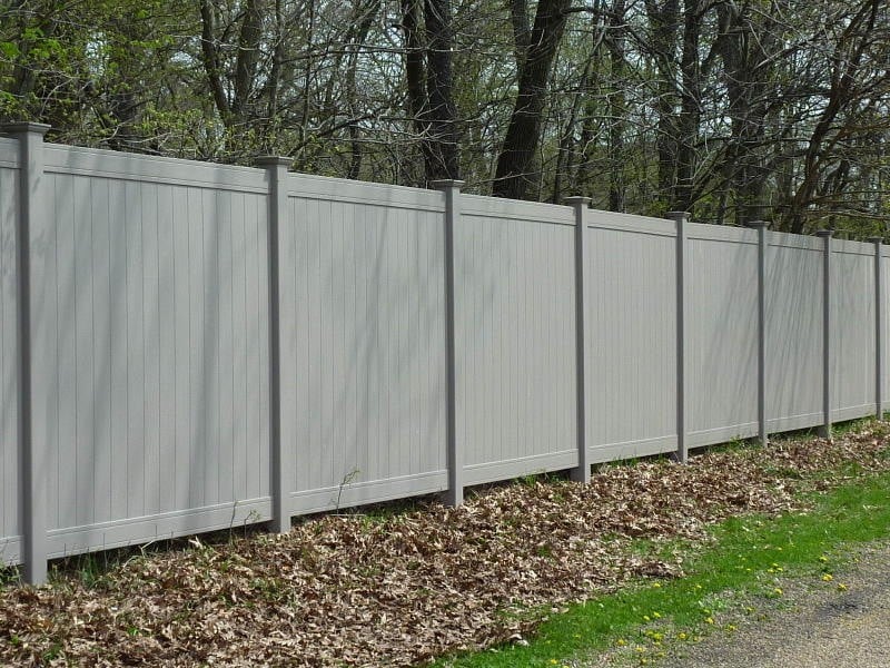 A light gray vinyl privacy fence borders a yard along a paved road.