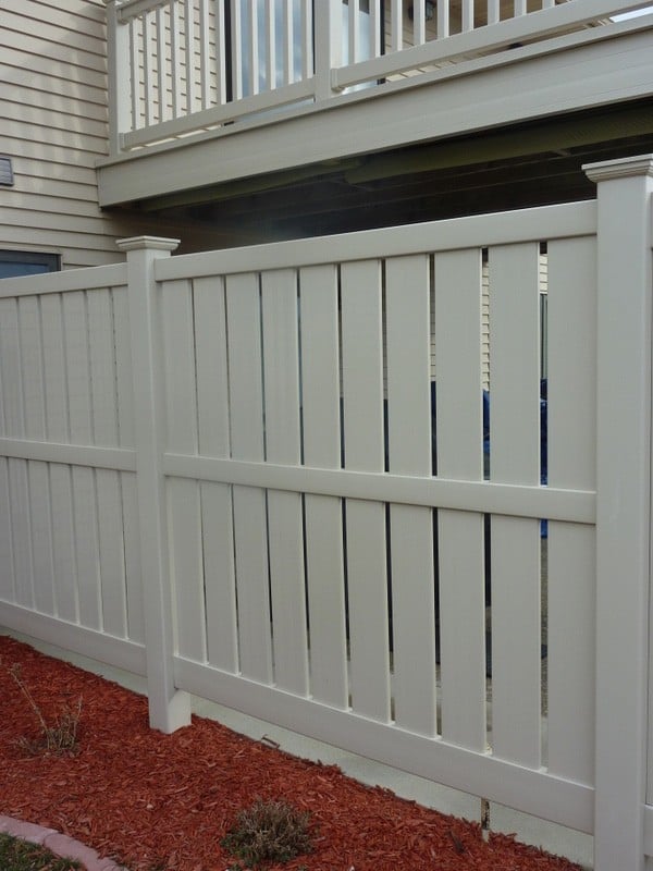 A tall beige fence in front of a house with matching siding, with red mulch and plants.