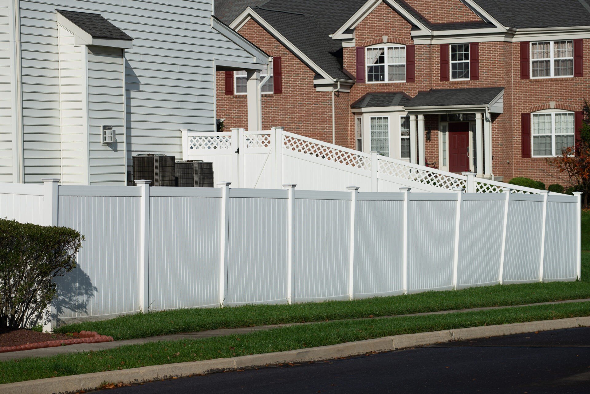 A tall white vinyl privacy fence bordering the lawn of a home with white siding, with a large red brick house beyond it.